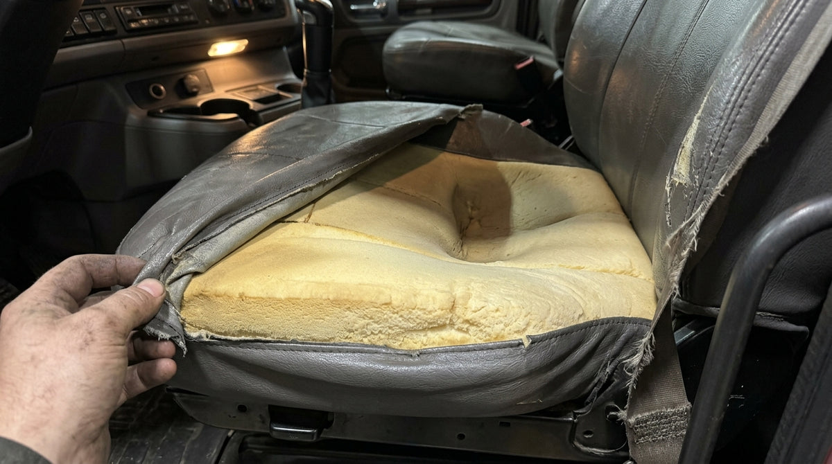 Documentary close-up, shot from above at a slight angle: the driver’s seat of a working truck with the cover pulled back, showing a distinct compressed hollow in the foam where the tailbone lands. Surrounding foam still full height. Worn vinyl and cab light, unstyled — the point is that this is an ordinary seat, not a ruined one.