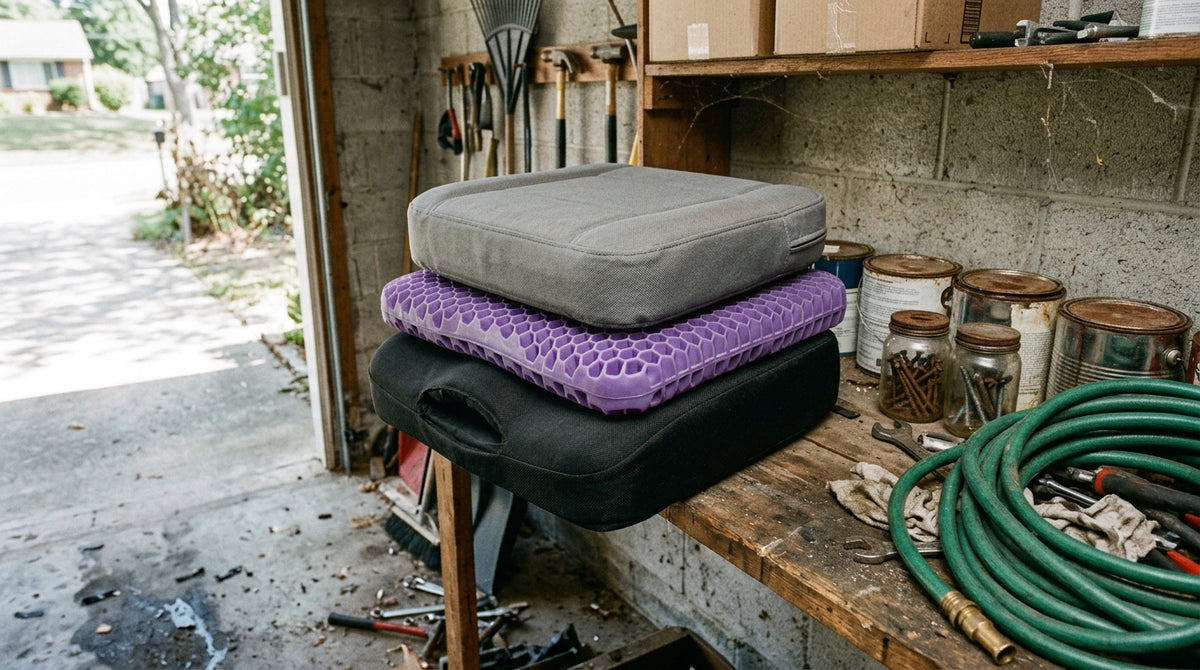 Documentary photograph in a suburban garage, natural light from an open door. Three used seat cushions stacked on a shelf beside paint tins and a coiled hose — one black foam pad with a visibly compressed hollow in the centre, one purple honeycomb gel grid slightly warped, one thick grey "trucker" pad. Dusty, unstyled, plainly abandoned. Shot slightly from above, as if by someone who just walked in and looked at them. No product, no text.