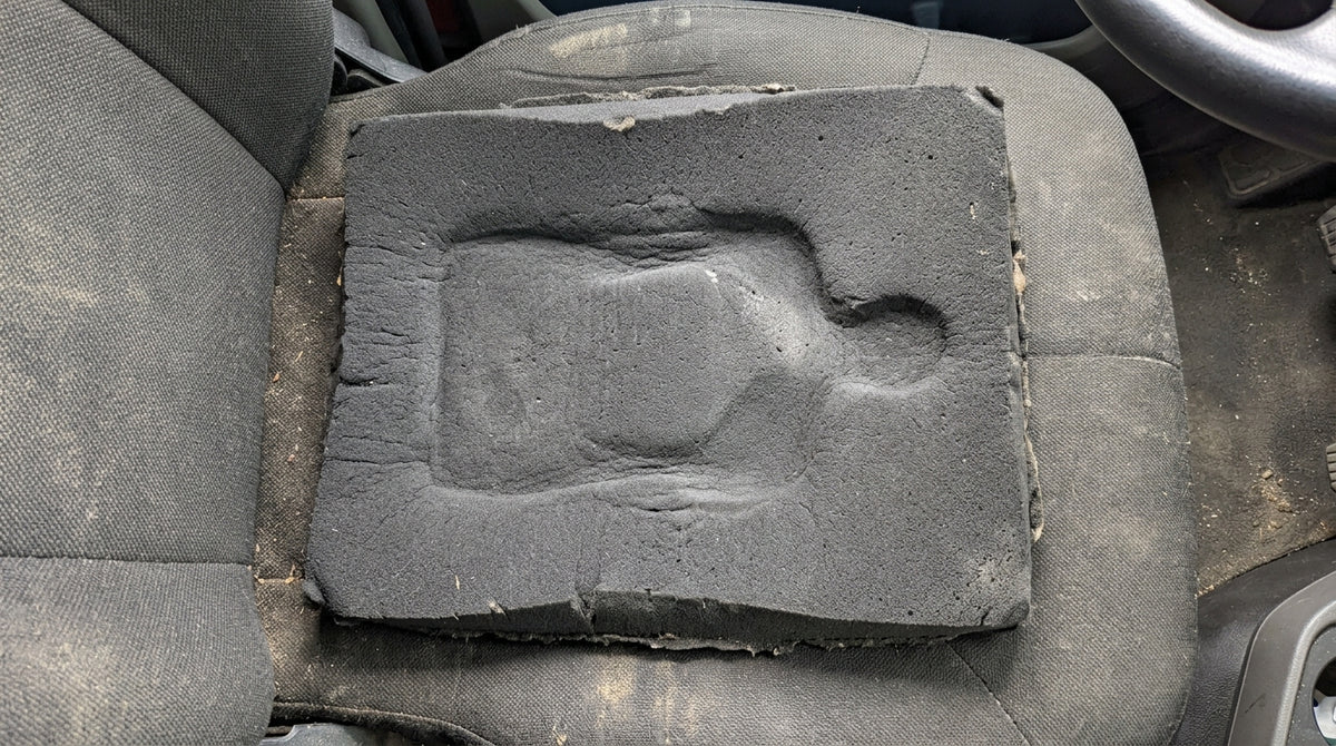 Overhead shot of a cheap black foam seat pad lying on a truck seat, visibly pancaked — a body-shaped dip compressed into the middle that has not come back. The pad is thinner at the dip than at the edges. Flat cab light, slightly grubby fabric. Evidence, not product photography.