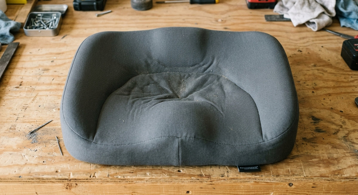A thick grey contoured seat pad, expensive-looking, photographed from above on a workbench. The centre has a clear compressed valley where the edges and corners are still full height. A brand label visible but not legible. The point of the frame is that the one part that mattered is the part that failed.